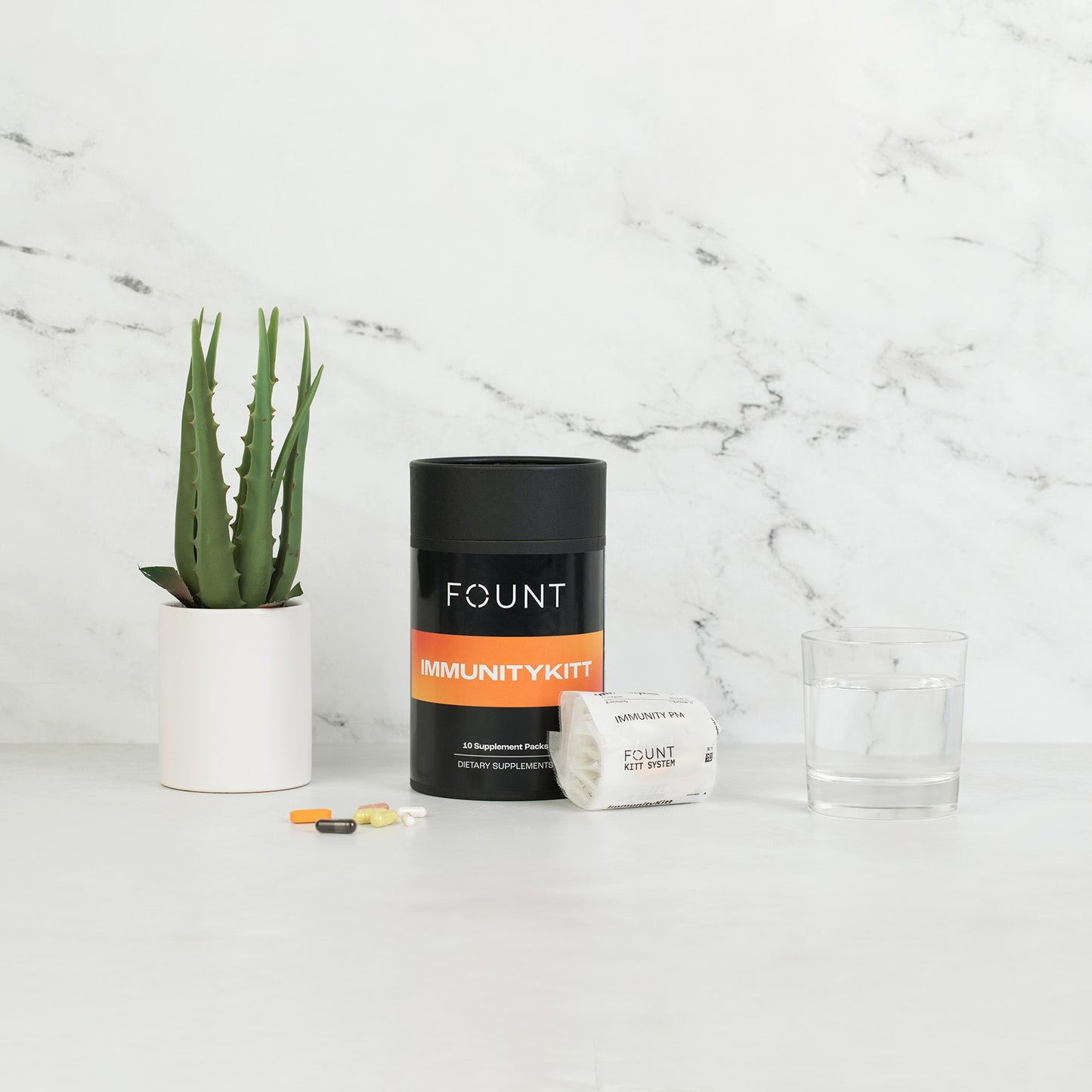 A container labeled IMMUNITYKITT by KITTS rests on a white surface with various pills, an open packet, a glass of water, and a small potted plant nearby. Set against a marble backdrop, this scene signifies not only immune support but also enhanced energy and mood.