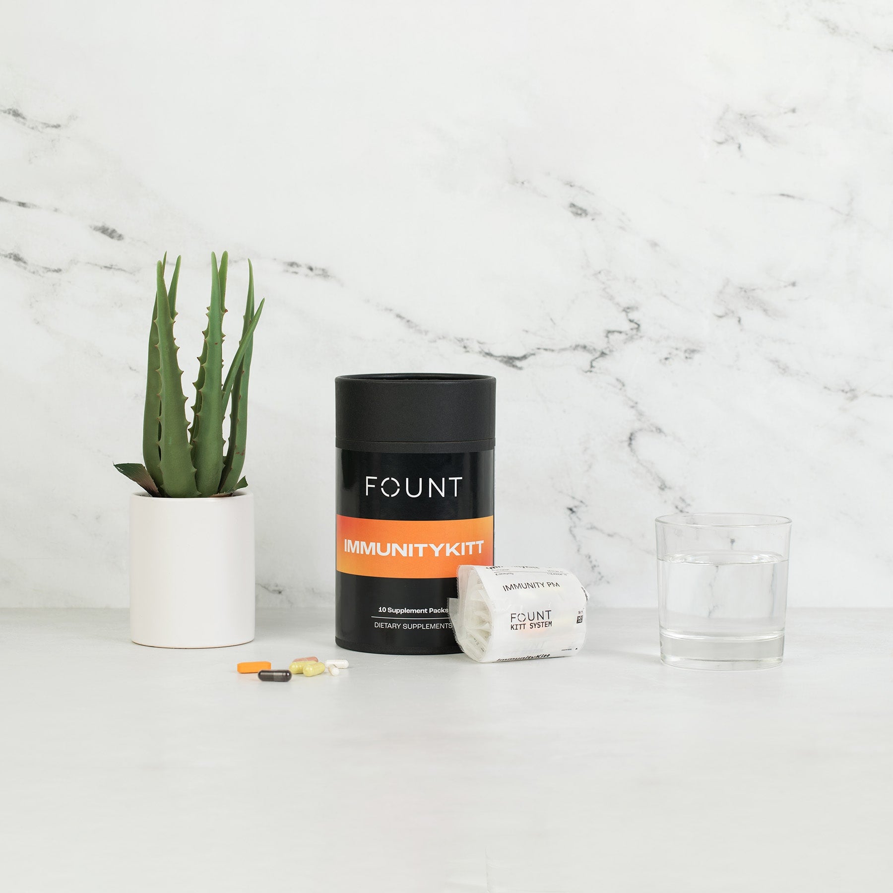 A container labeled IMMUNITYKITT by KITTS rests on a white surface with various pills, an open packet, a glass of water, and a small potted plant nearby. Set against a marble backdrop, this scene signifies not only immune support but also enhanced energy and mood.